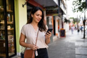 Mulher sorrindo enquanto olha para o celular em uma calçada urbana.